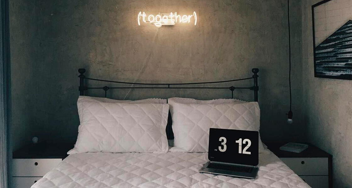 um quarto com roupas de cama brancas e parede cinza, criados-mudo brancos e uma placa luminosa escrito "(together)" acima da cama.