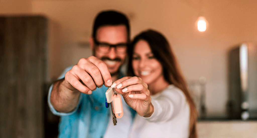 casal segura junto a chave do novo apartamento, enquanto sorriem e olham para a câmera